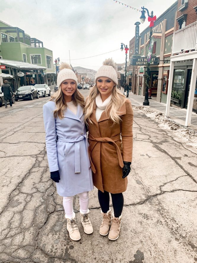 two blonde girls with blue and tan trench coats and winter clothing in the snowy town of park city, utah